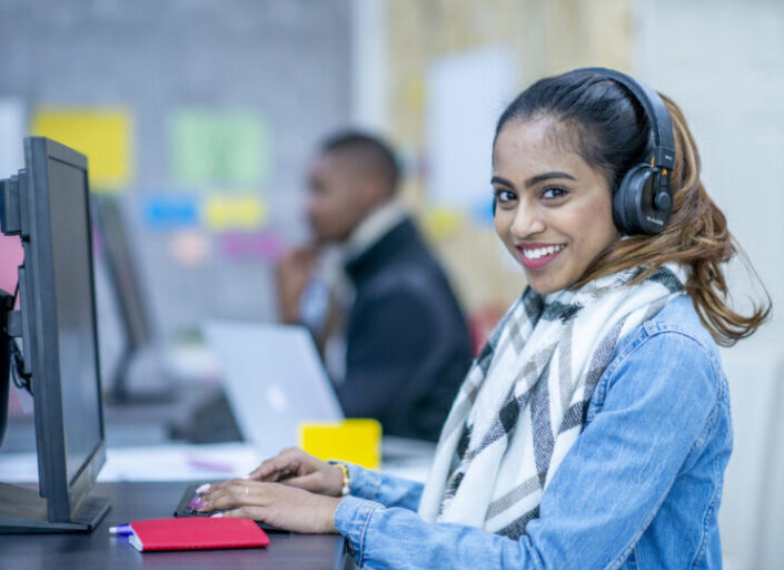 A young woman in casual clothing is sitting on a computer and working. She is wearing a set of black headphones.