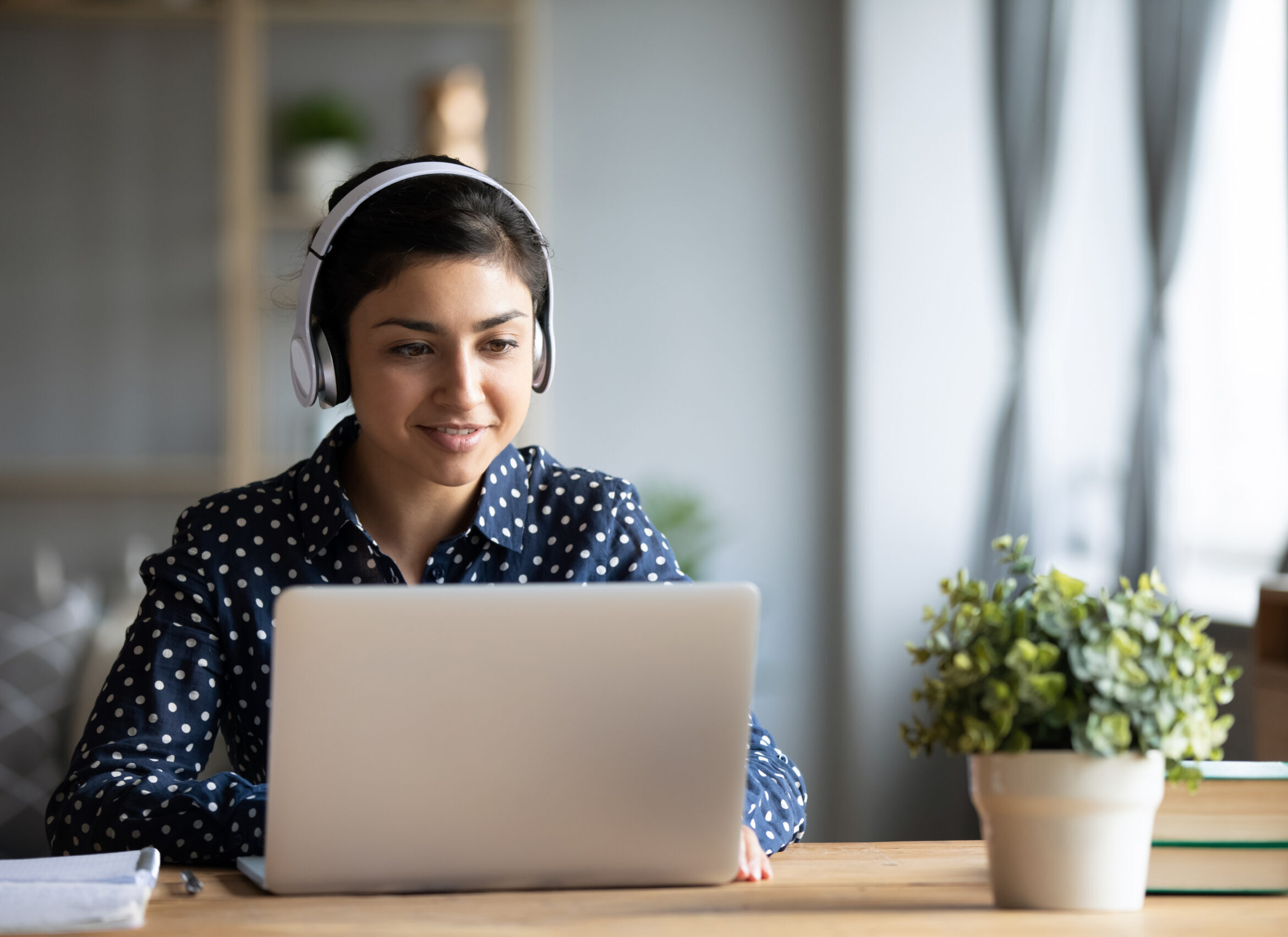 Millennial Indian girl in wireless headphones sit at desk at home working on modern laptop, young ethnic woman in earphones browsing Internet shopping online or studying on computer in living room